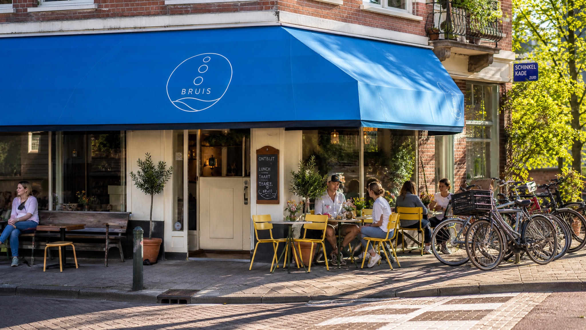 Het zonnige hoekpand van Bruis met kobaltblauwe luifel en het bubbel-logo, gele terrasstoelen