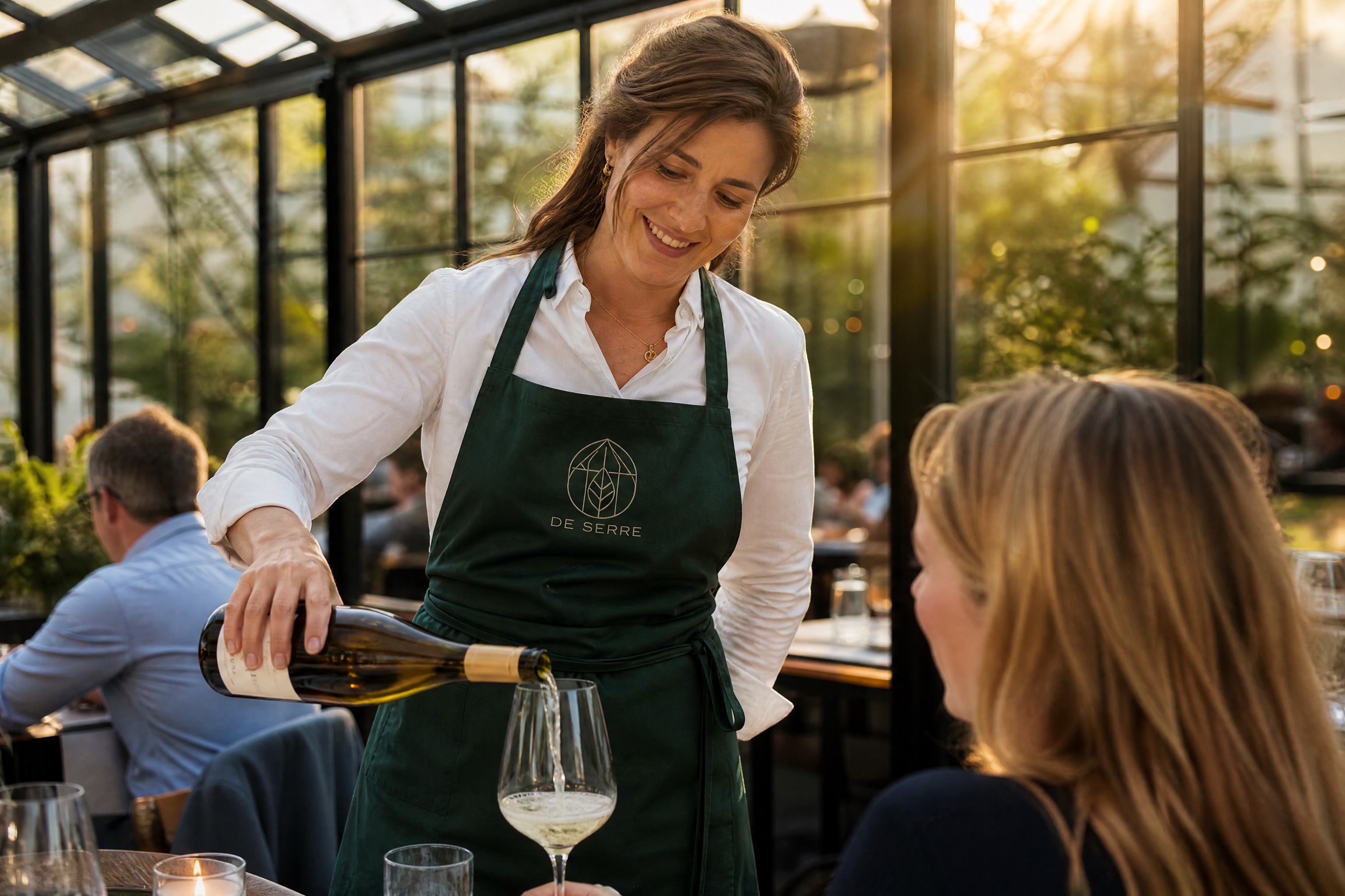 De sommelier van De Serre schenkt wijn in de kas, het monogram in ecru garen op haar schort